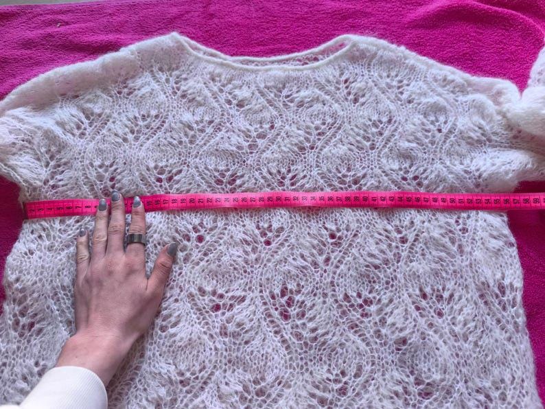 May include: A cream-colored, open-weave sweater with a round neckline. A pink measuring tape is stretched across the chest of the sweater. The sweater's width is being measured. The sweater has a delicate, lacy pattern.