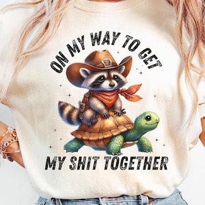 May include: Cream-coloured t-shirt with a raccoon in a cowboy hat riding a turtle. The raccoon wears a red bandana, and the turtle has a green shell. The text "ON MY WAY TO GET MY SHIT TOGETHER" is printed on the shirt in black lettering.