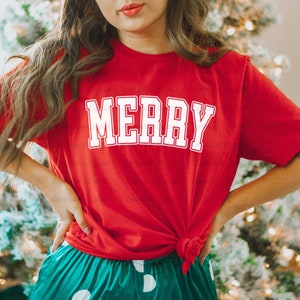 May include: Red t-shirt with white text that says "MERRY" in a varsity style font. The shirt is tied at the waist and worn with green and white polka dot pants.
