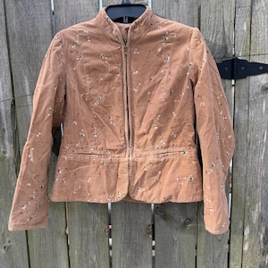 May include: A tan corduroy jacket with a mandarin collar and a full-length zipper. The jacket is adorned with embroidered floral details in white, green, and brown. It has two front pockets and is hanging on a black hanger against a wooden fence.