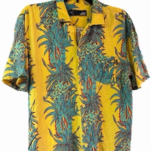 May include: A short-sleeve button-down shirt in bright yellow featuring a repeating vertical pattern of teal and orange pineapples. The shirt has a standard collar, a single chest pocket on the left side, and a black label at the neck marked with an M.