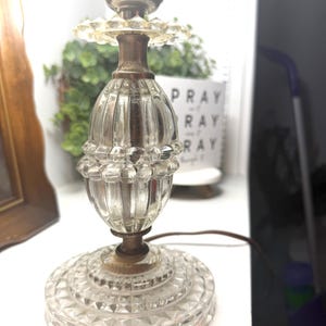 May include: A vintage table lamp base featuring a clear, pressed glass body with a ribbed, oval-shaped center section and a circular, faceted base. The lamp includes a metallic socket at the top and a brown electrical cord extending from the side.