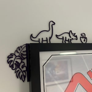 以下が含まれることがあります： 黒い額縁の上に、紫色の恐竜、紫色のトリケラトプス、紫色の恐竜の卵が置かれている。額縁の角には、紫色のモンステラの葉のデザインが施されている。