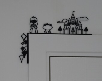 May include: Black decorative corner piece featuring a princess, frog, castle, and diamond shapes. The princess wears a crown and dress. The castle has multiple turrets. The frog is smiling. The corner piece is mounted on a white door.