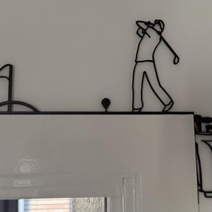 May include: Black metal golf-themed door topper featuring a golfer swinging a club, a golf ball, a flag on a green, and a golf bag with clubs. The design is mounted on a white door.