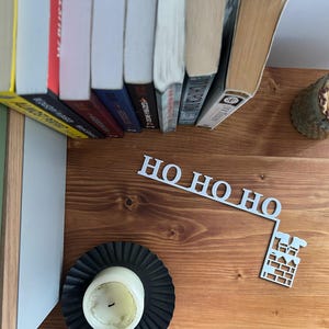 May include: A wooden shelf with books, a candle, and a decorative metal sign that says "HO HO HO" with a chimney design. A small container of matches sits on the shelf.