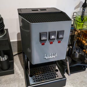 以下が含まれることがあります： グレーのGaggia Classicエスプレッソマシン。3つの黒いコントロールスイッチとステンレス製のドリップトレイが付いています。マシンにはGaggia Classicのロゴがあります。