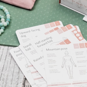 A set of white yoga pose cards with illustrations and instructions. The cards are laid out on a green yoga mat with a turquoise beaded necklace and a few small stones.