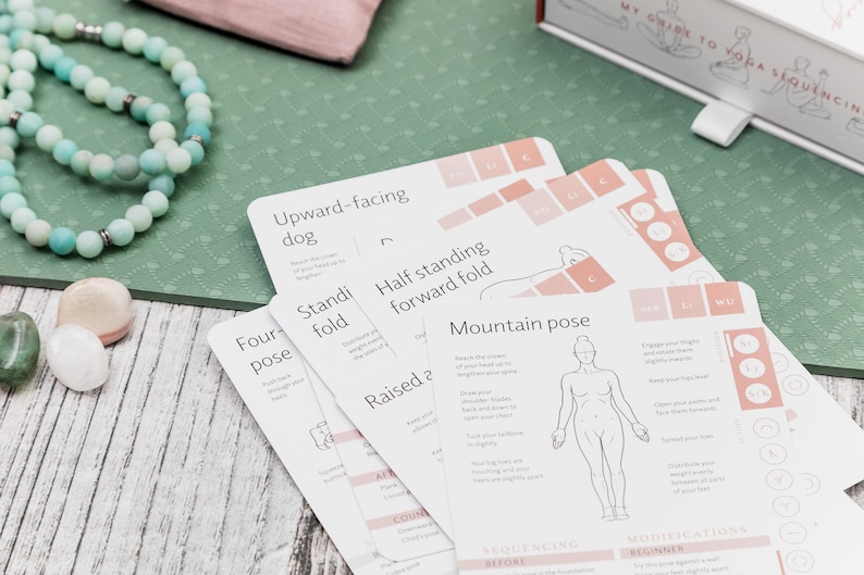 A set of white yoga pose cards with illustrations and instructions. The cards are laid out on a green yoga mat with a turquoise beaded necklace and a few small stones.