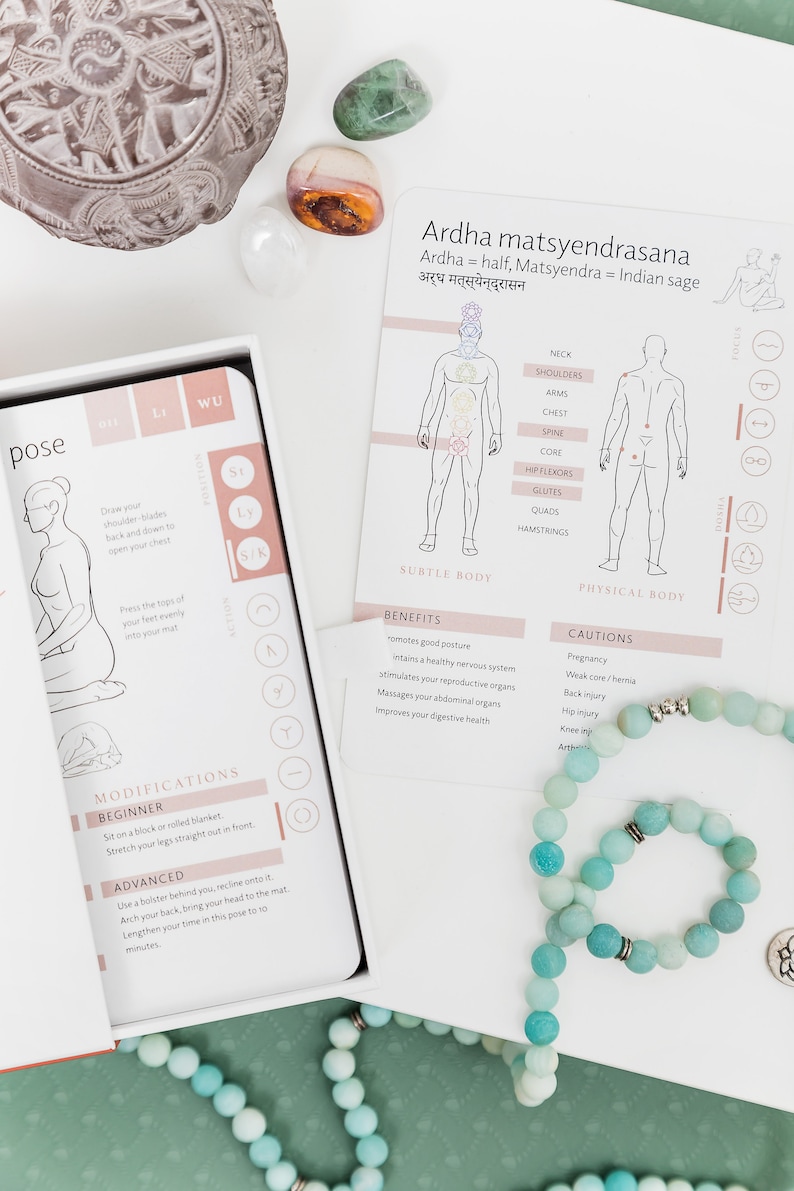 A white card with the name of a yoga pose, Ardha Matsyendrasana, and a description of the pose's benefits and cautions. The card is surrounded by a teal blue mala necklace and a few colourful stones.