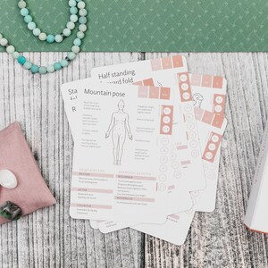 A set of yoga pose cards with illustrations and instructions. The cards are on a wooden surface with a pink pouch containing crystals and a string of beads.