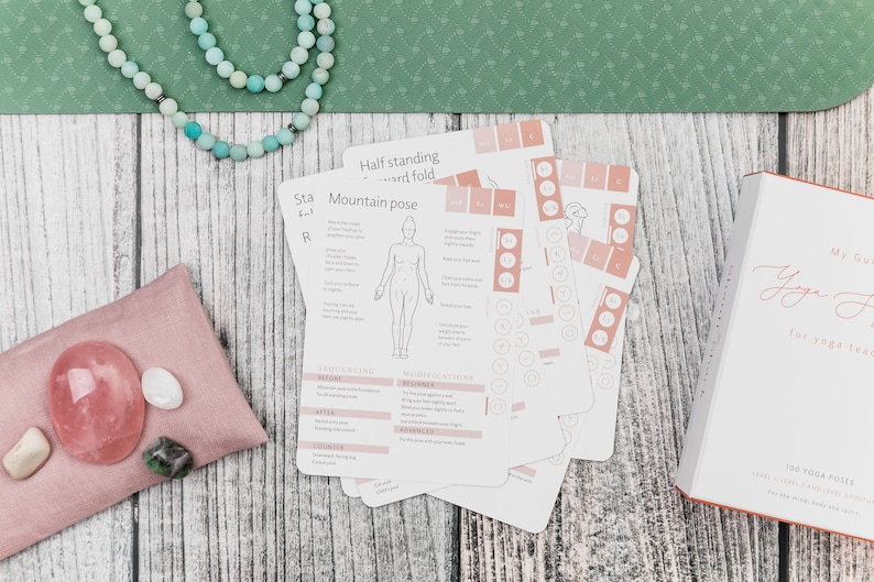 A set of yoga pose cards with illustrations and instructions. The cards are on a wooden surface with a pink pouch containing crystals and a string of beads.