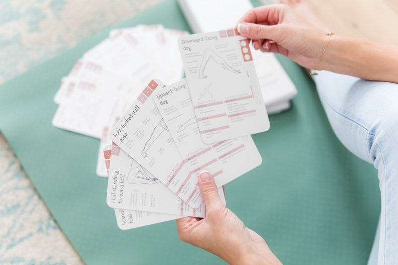 A set of white yoga pose cards with pink and brown accents. The cards feature illustrations of yoga poses and the names of the poses, such as "Downward-facing dog" and "Four-limbed staff pose".