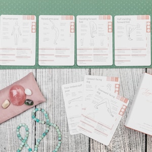 A set of yoga pose cards with illustrations and instructions. The cards are laid out on a green yoga mat with a pink eye pillow, a string of turquoise beads, and a book titled "My Guide to Yoga Journey" on a wooden surface.