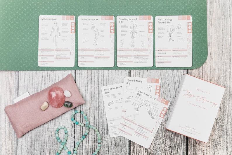 A set of yoga pose cards with illustrations and instructions. The cards are laid out on a green yoga mat with a pink eye pillow, a string of turquoise beads, and a book titled "My Guide to Yoga Journey" on a wooden surface.
