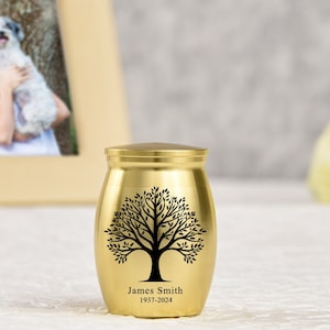 Könnte beinhalten: Goldene Metallurne mit einem schwarzen Baumdesign und der Inschrift "James Smith 1937-2024".