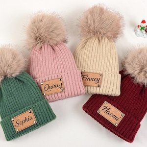 Könnte beinhalten: Vier gerippte Strickmützen in Grün, Rosa, Beige und Burgunderrot, jede mit einem flauschigen Bommel. Jede Mütze hat ein rechteckiges Kunstleder-Patch mit einem Namen: Sophia, Quincy, Finney und Naomi.