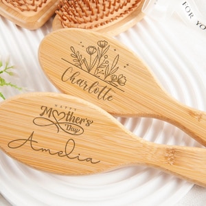 Könnte beinhalten: Zwei Holzhaarbürsten mit personalisierten Gravuren. Eine Bürste sagt "Happy Mother's Day Amelia" und die andere sagt "Charlotte" mit einem floralen Design.