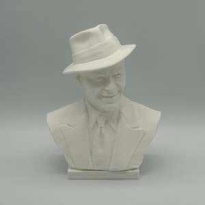 Pode incluir: Busto de escultura branca de uma pessoa usando um fedora e um paletó. A escultura está sobre uma base branca.