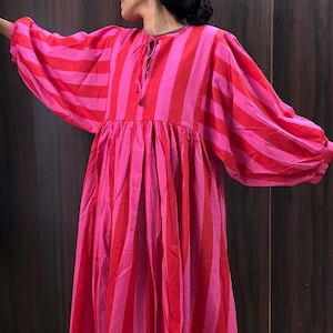 Puede incluir: Un vestido largo y fluido con rayas verticales en tonos rosa y rojo. El vestido presenta mangas abullonadas voluminosas y una cintura fruncida. El escote tiene un cierre de lazo. El vestido está hecho de una tela ligera.