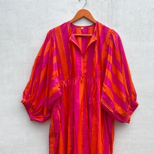 May include: A vibrant, oversized dress with vertical stripes in bright pink and orange. The dress features long, voluminous sleeves and a collared neckline. It hangs on a wooden hanger against a neutral background.