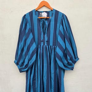 Op de afbeelding: Een blauw en marine gestreepte jurk met een ronde hals en strikdetail. De jurk heeft driekwart mouwen en een gerimpelde rok. De jurk hangt aan een houten hanger.