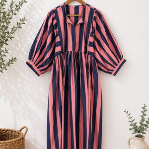 May include: A midi-length dress with vertical stripes in navy blue and coral pink. The dress features a collared neckline, three-quarter sleeves, and a gathered waist. It hangs on a wooden hanger against a white wall.