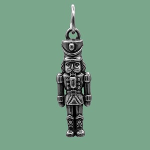 May include: A silver-colored charm in the shape of a nutcracker soldier. The charm has a loop at the top for hanging and features detailed facial features, a hat, and boots with skull and crossbones. The background is a solid green color.