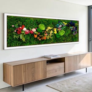 May include: A rectangular wall art piece featuring a vibrant arrangement of colorful flowers and greenery set against a backdrop of lush green moss. The artwork is framed in a clean white border, creating a striking contrast. The piece is mounted above a wooden cabinet.