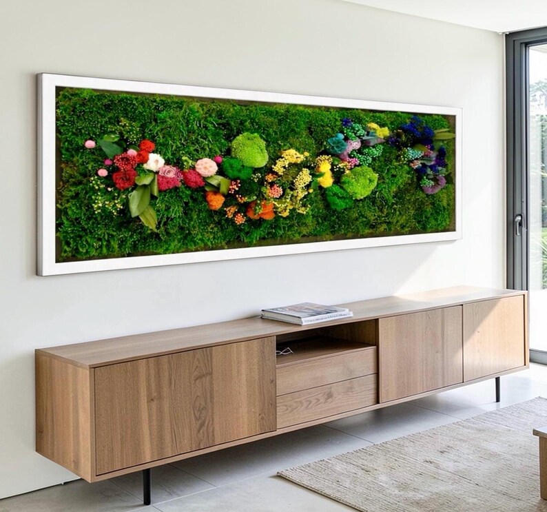 May include: A rectangular wall art piece featuring a vibrant arrangement of colorful flowers and greenery set against a backdrop of lush green moss. The artwork is framed in a clean white border, creating a striking contrast. The piece is mounted above a wooden cabinet.