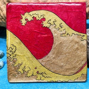 May include: A square coaster with a red background and a gold wave design. The wave is stylised and has a textured look.