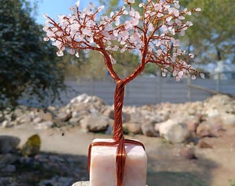 Pode incluir: Uma árvore de arame envolto em quartzo rosa com uma base de quartzo rosa. A árvore é feita de arame de cobre e tem uma forma natural e orgânica. A base é um bloco quadrado de quartzo rosa.