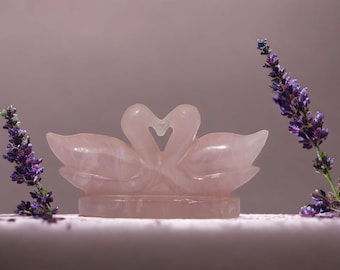 Op de afbeelding: Een roze rozenkwarts sculptuur van twee zwanen met hun nekjes in elkaar gevlochten in de vorm van een hart. Het beeld staat op een wit oppervlak met lavendelbloemen aan beide kanten.