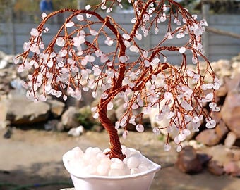Pode incluir: Uma árvore envolta em arame com cristais de quartzo rosa, em um vaso branco em forma de coração, sobre uma superfície de pedra cinza.