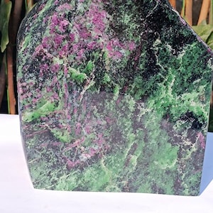 Può includere: Una grande scultura in pietra lucida dalla forma unica. La pietra presenta un sorprendente motivo di tonalità verdi, nere e rosa. La scultura è posta su una superficie bianca e uno sfondo di verde.