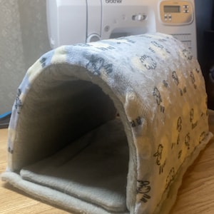 Può includere: Un letto per animali domestici a forma di tunnel, con motivo grigio e bianco. Il letto è realizzato in tessuto morbido e peluche e ha un fondo piatto.