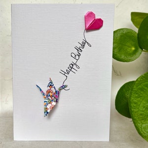 May include: A white greeting card featuring a colorful floral origami crane at the bottom and a metallic pink origami heart at the top, connected by a black cursive line of text that reads Happy Birthday in a vertical orientation.
