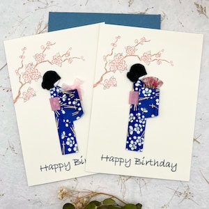 May include: Two cream-coloured birthday cards with a Japanese theme. Each card features a blue kimono with white floral patterns, a black hairstyle, and a pink bow or fan. Cherry blossom branches are painted above the figures, and the words "Happy Birthday" are written below.