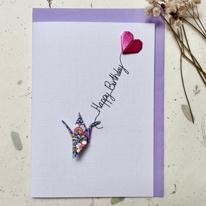 May include: A white greeting card with a textured surface featuring a colorful floral origami crane and a small metallic pink origami heart. The text Happy Birthday is written in black script, connecting the crane to the heart shape.