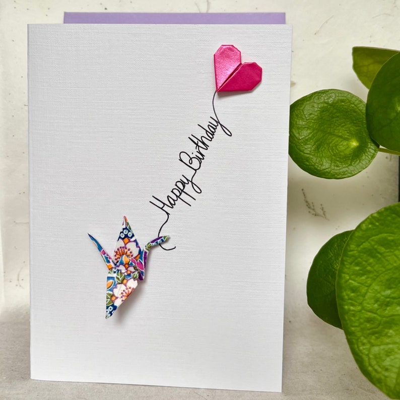 May include: A white greeting card featuring a folded pink origami heart at the top and a colorful floral patterned origami crane at the bottom. The text Happy Birthday is written in black cursive script, connecting the heart and the crane.