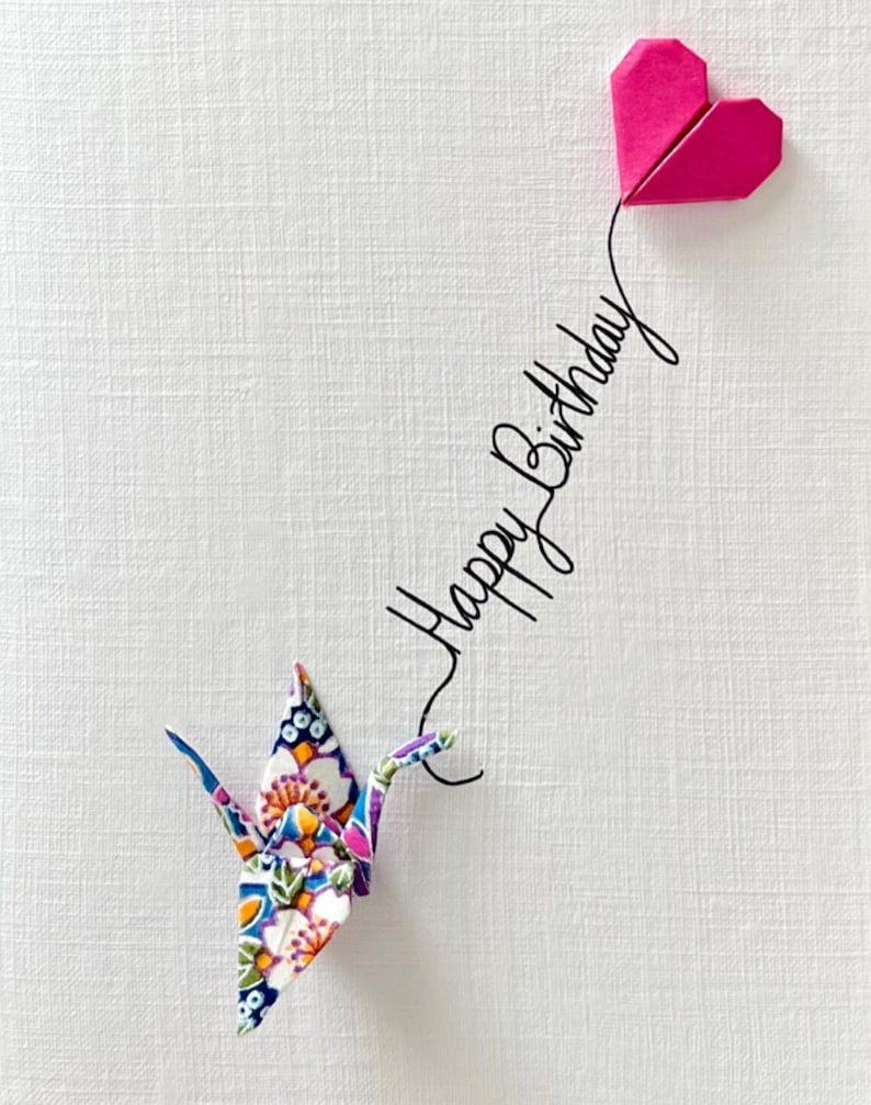 May include: A white textured greeting card featuring a colorful floral origami crane connected by a black line to a pink origami heart. The text Happy Birthday is written in black cursive script along the line connecting the crane and the heart.