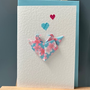 Handgemachte Grußkarte im Origami-Design mit verliebten Kranichen in einer Herzformation und türkisen Blüten und Herzen