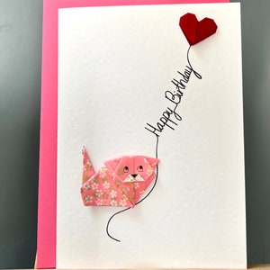 Carte de voeux faite main en origami avec un chat, des fleurs blanches roses et un ballon coeur rouge « Happy Birthday »