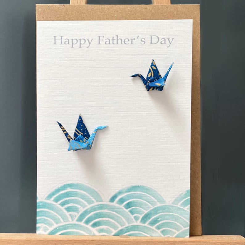 Op de afbeelding: Een witte kaart met de tekst "Happy Father's Day" en twee blauwe origami-kranen. De kaart heeft een blauw en groen golfpatroon aan de onderkant.