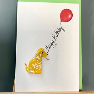 Carte de voeux en origami chien assis jaune, fleurs blanches et ballon rouge « Happy Birthday »