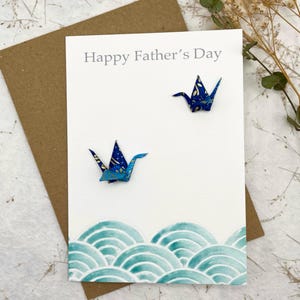 Op de afbeelding: Een witte kaart met de tekst "Happy Father's Day" in grijs gedrukt. Twee blauwe origami-kranen zijn op de kaart geplakt. De onderkant van de kaart heeft een blauw en wit golfpatroon.
