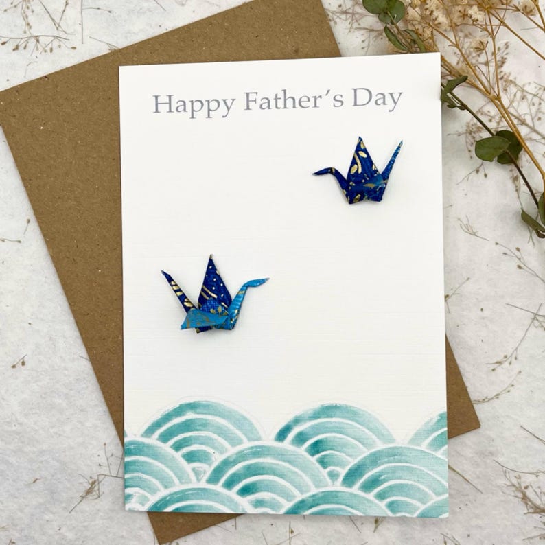 Op de afbeelding: Een witte kaart met de tekst "Happy Father's Day" in grijs gedrukt. Twee blauwe origami-kranen zijn op de kaart geplakt. De onderkant van de kaart heeft een blauw en wit golfpatroon.