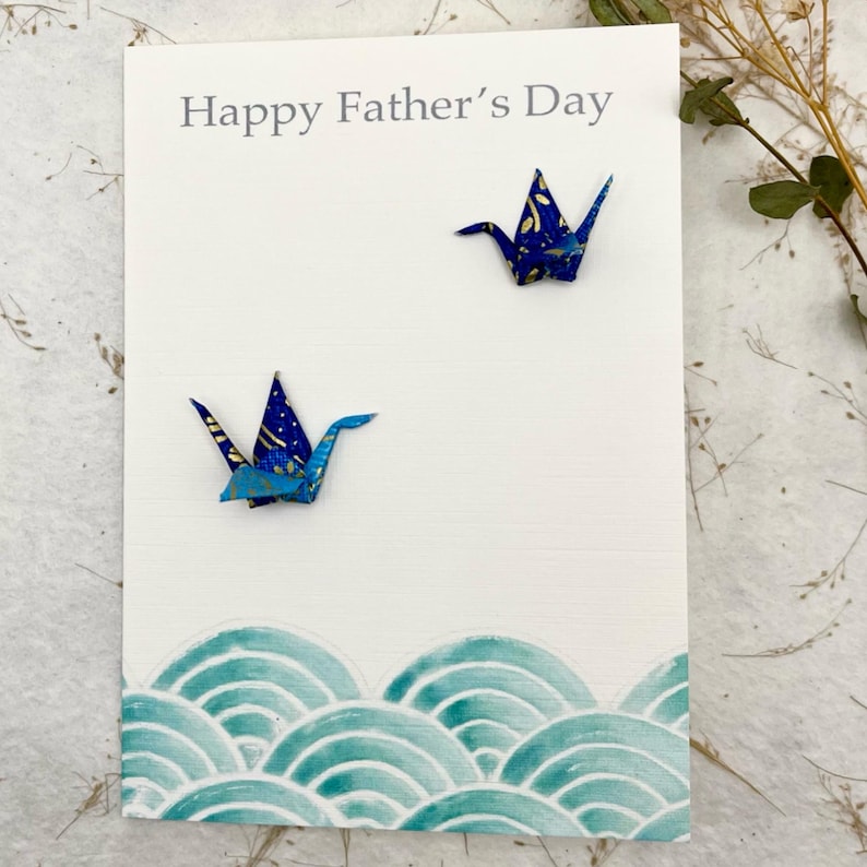 Op de afbeelding: Een witte kaart met de tekst "Happy Father's Day" in grijs gedrukt. Twee blauwe origami-kranen zijn aan de kaart bevestigd. De onderkant van de kaart heeft een blauw en groen golfpatroon.