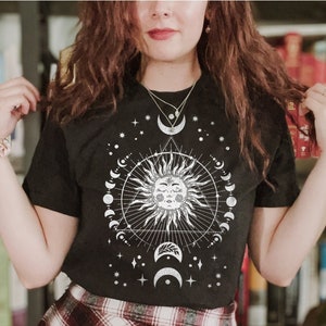 K&ouml;nnte beinhalten: Schwarzes T-Shirt mit einem wei&szlig;en Sonnen- und Mondmotiv. Das Motiv zeigt eine Sonne mit einem Gesicht und einen Kreis aus Monden darum herum. Das Shirt wird mit einem rot-wei&szlig; karierten Rock kombiniert.