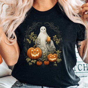 May include: Black t-shirt featuring a Halloween-themed graphic. The design includes a white ghost holding a candle, surrounded by pumpkins and foliage. The shirt is a crew neck and the color is black.